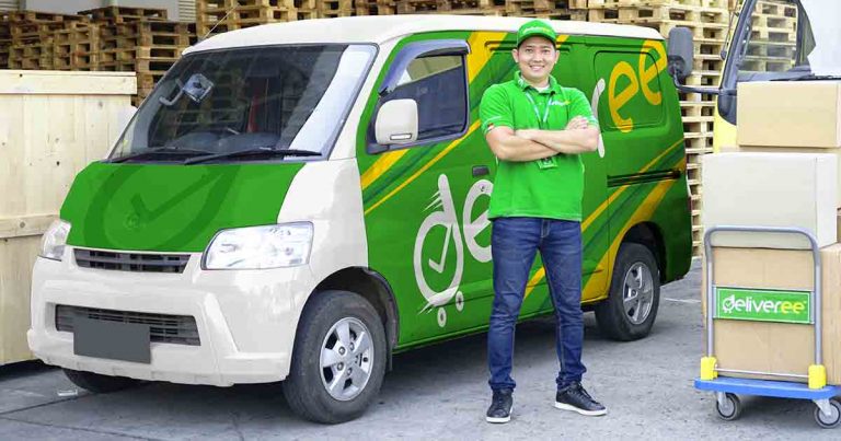 Seorang driver tersenyum mengenakan seragam hijau berdiri di samping van pengiriman barang Deliveree berwarna hijau dan kuning dengan beberapa kotak di troli pengiriman.