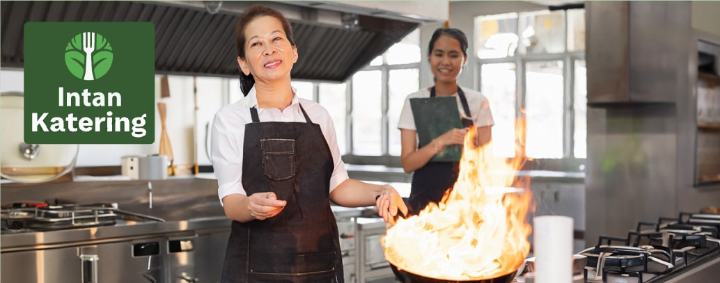 Seorang wanita sedang memasak makanan dengan api besar dan diperhatikan oleh asisten wanitanya