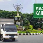 Truk box Deliveree yang sedang berada di dekat bundaran dengan latar belakang pepohonan dan air mancur hias. Sebuah papan tanda berwarna hijau di samping truk bertuliskan "Jasa Sewa Truk Karawang," yang menunjukkan layanan sewa truk di Karawang.