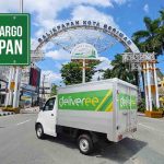 Mobil truk Deliveree putih melaju di jalan Balikpapan, dengan gerbang 'Balikpapan Kota Beriman' dan papan 'Ekspedisi Cargo Balikpapan' di latar belakang.