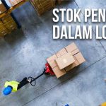 Pekerja di gudang memindahkan stok pengaman dengan alat dorong di antara rak logistik.