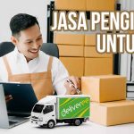 Pengusaha UKM tersenyum sambil dikelilingi kotak paket dengan miniatur truk Deliveree di depannya sebagai ilustrasi jasa pengiriman yang digunakan.