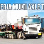 Truk multi axle trailer mengangkut muatan berat yang ditutup terpal di area logistik dekat kontainer.