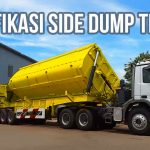 *Truk trailer side dump berwarna kuning sedang parkir di depan bangunan dengan tulisan 'Spesifikasi Side Dump Trailer'.